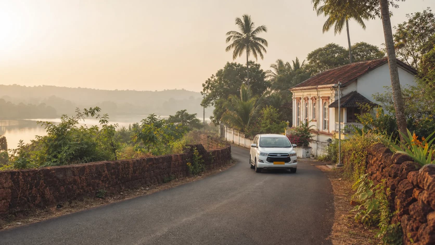 A white Goa taxi on a quiet riverside road beside laterite walls and a heritage home at sunrise